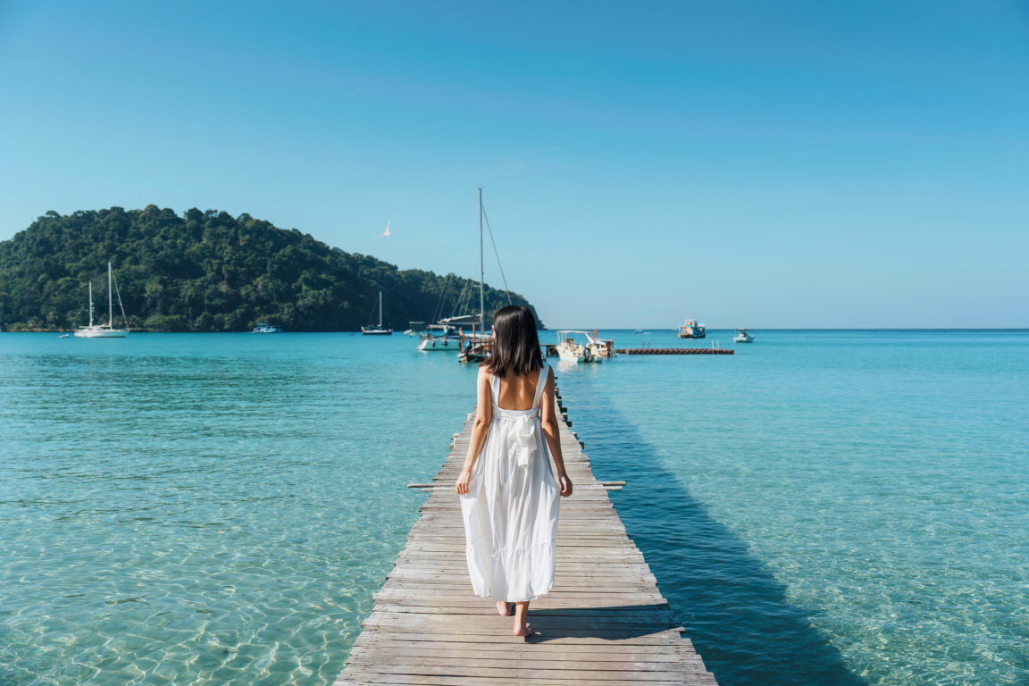 Mujer asiática joven en el vestido blanco que camina en el embarcadero de madera en mar tropical en día soleado